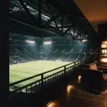 A dramatically lit sports arena with warm neon lights and a slot machine visible in a nearby lounge