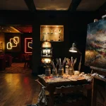 Paint-covered easel and richly textured painting in a dark gallery with a softly lit casino lounge behind it