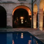 Azure-tiled courtyard pool under cinematic evening light, with a warm neon gaming lounge and slot machine beyond the arches