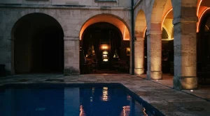 Azure-tiled courtyard pool under cinematic evening light, with a warm neon gaming lounge and slot machine beyond the arches