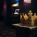 A golden crown built from chunky arcade-style blocks on a dark plinth, with a softly lit arcade cabinet behind it