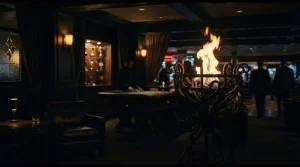 A bright flame rises from a metal brazier beside a dark gaming lounge with a softly lit roulette table and trophy shelf
