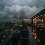 Billowing clouds above a rooftop lounge at dusk, with a vintage arcade cabinet glowing in warm neon light.