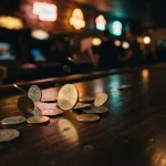 Gold and silver coins spilling onto a dark wooden counter beside a softly glowing arcade cabinet in a neon-lit gaming lounge