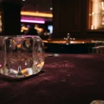 A glass gaming die on a dark felt table under warm neon light, with a roulette wheel and prize shelf behind