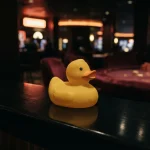 A yellow rubber duck on a glossy dark counter with a warmly lit casino game table in the background
