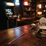 Two crossed fencing foils beside protective masks on a dark table, with a warmly lit arcade cabinet in the background