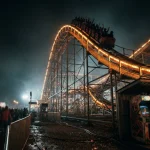 An illuminated fairground rollercoaster rises above a dark night scene, with a small vintage arcade cabinet nearby.