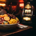 A bowl of ripe tropical fruit on a dark polished table, with warm neon light and a softly glowing slot machine behind it