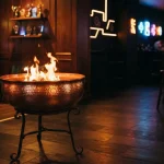Copper brazier with lively orange flames in a dark neon-lit gaming lounge, with arcade machines and a trophy shelf behind it