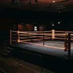 Empty boxing ring under amber arena lights, with a glowing slot cabinet inside a shadowy lounge beyond