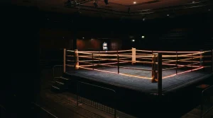 Empty boxing ring under amber arena lights, with a glowing slot cabinet inside a shadowy lounge beyond