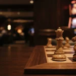 An ivory chess queen and knight on a polished board in a dim lounge, with a softly lit slot machine behind them.