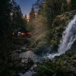 A wild forest waterfall beside a dark timber gaming lodge with a warmly lit arcade cabinet in the distance