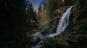 A wild forest waterfall beside a dark timber gaming lodge with a warmly lit arcade cabinet in the distance