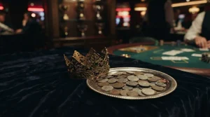 An ornate brass crown beside a silver coin tray on dark velvet, with a warmly lit casino card table behind