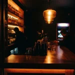 A glowing late-night bar counter with glassware and a softly lit arcade cabinet in the corner