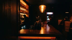 A glowing late-night bar counter with glassware and a softly lit arcade cabinet in the corner
