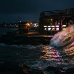 A rolling ocean wave illuminated by warm neon beside a small seaside arcade at night, under cinematic dark skies
