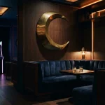 Crescent moon sculpture above a dark, warmly lit lounge with an arcade cabinet glowing in the background