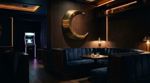 Crescent moon sculpture above a dark, warmly lit lounge with an arcade cabinet glowing in the background