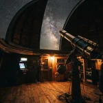 Open observatory dome beneath a star-filled sky with a warmly illuminated arcade cabinet in the background