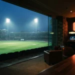 Floodlit cricket stadium at dusk with a small arcade cabinet and trophy shelf in a warm neon-lit lounge