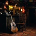 An acoustic guitar and empty wooden chair in a warmly lit Mexican cantina, with a discreet slot machine in the background