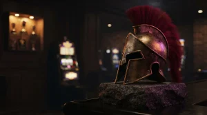 Weathered bronze Spartan helmet on a dark stone plinth with a softly glowing arcade machine in the background