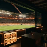 A floodlit football stadium beyond a dark games lounge with a vintage arcade cabinet and a trophy shelf