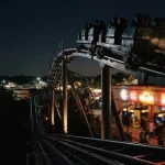A dramatic nighttime rollercoaster cresting a steep track, with glowing arcade machines in the distance