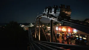 A dramatic nighttime rollercoaster cresting a steep track, with glowing arcade machines in the distance