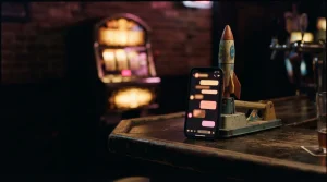 A glowing phone beside a miniature rocket arcade game and a softly lit gaming cabinet in a dark neon lounge