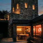 An illuminated stone watchtower above a dark arcade lounge with a softly glowing game table and warm neon lighting