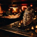 Carved ceremonial mask beside glowing candles in a moody night bar, with a softly lit gaming table in the background